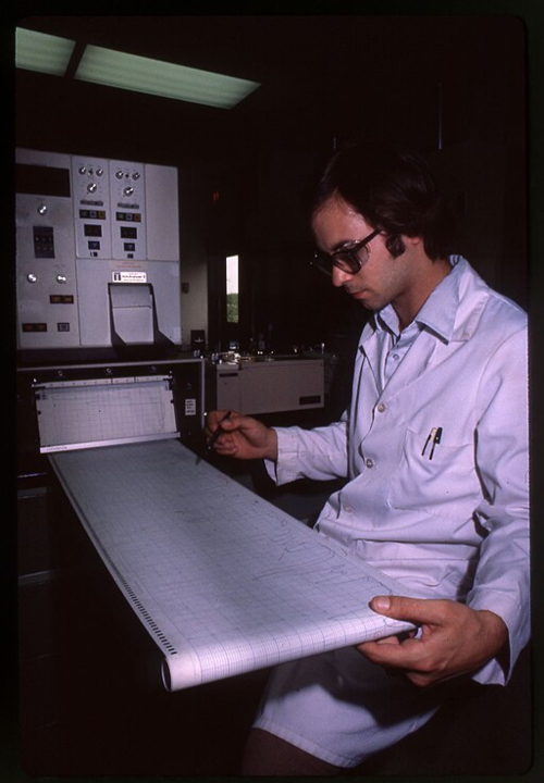 Male scientist wearing glasses and a white lab coat. Sitting next to a large computer and printer while holding a pen and studying a large printout on graph paper.